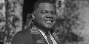 Picture of a young man smiling wearing a suit with Kente collegiate stole around his neck