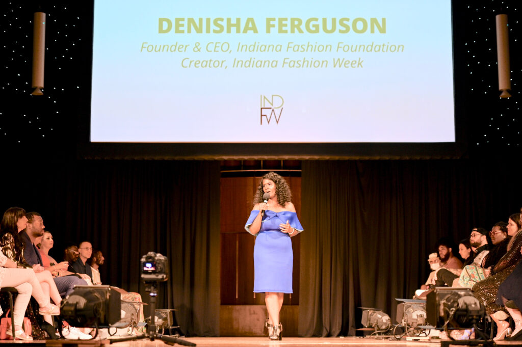 Woman wearing a off the shoulder blue dress at Indiana Fashion Week in front of a group of people